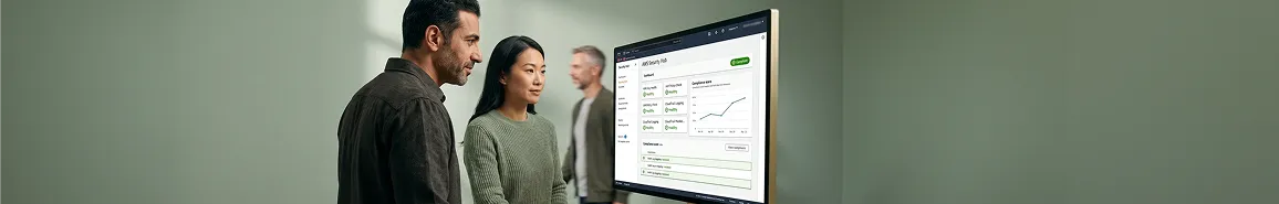 Compliance lead and engineer reviewing an all-green cloud security and compliance audit dashboard on a large monitor after migration in soft office side light Compliance lead and engineer reviewing an all-green cloud security and compliance audit dashboard on a large monitor after migration in soft office side light