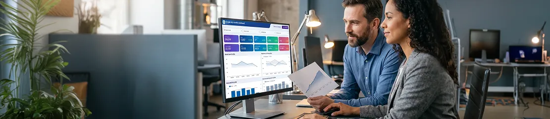 Two-person paid media team at adjacent desks reviewing Google Ads campaign performance improvement graphs, warm desk lamps, side angle Two-person paid media team at adjacent desks reviewing Google Ads campaign performance improvement graphs, warm desk lamps, side angle